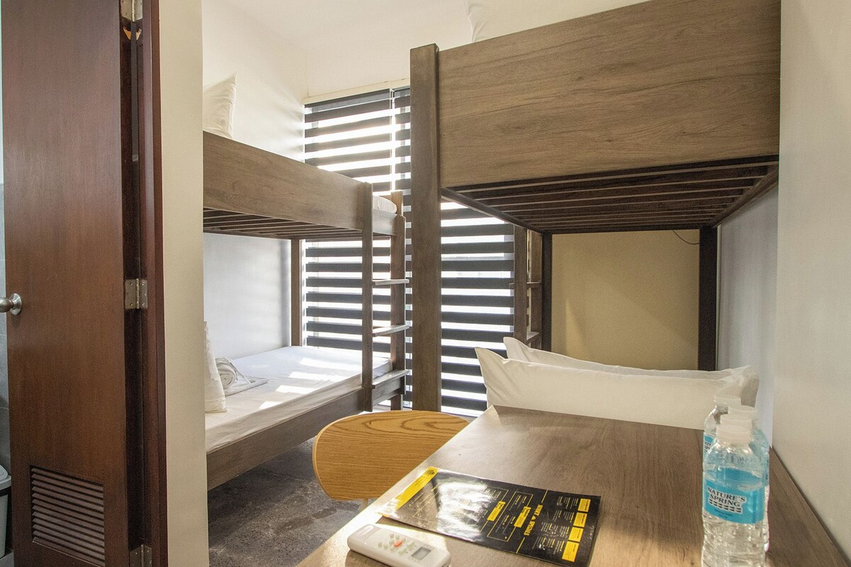 A compact room with bunk beds, a small table with a book and a bottle, a toilet, and a shower area.