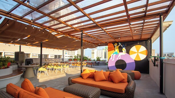 An outdoor seating area with wicker sofas and orange cushions, a large mural, and a view of the cityscape. - The Pad Co-living (Cebu City)