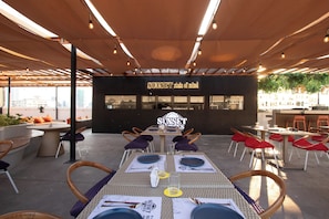 An outdoor dining area with tables and chairs, a sign reading "SUNSET state of mind", and a view of buildings in the background. - The Pad Co-living (Cebu City)