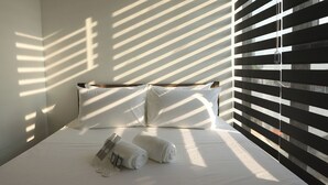 A neatly made bed with white linens and two rolled towels, positioned under a window with horizontal blinds casting shadows on the wall. - The Pad Co-living (Cebu City)