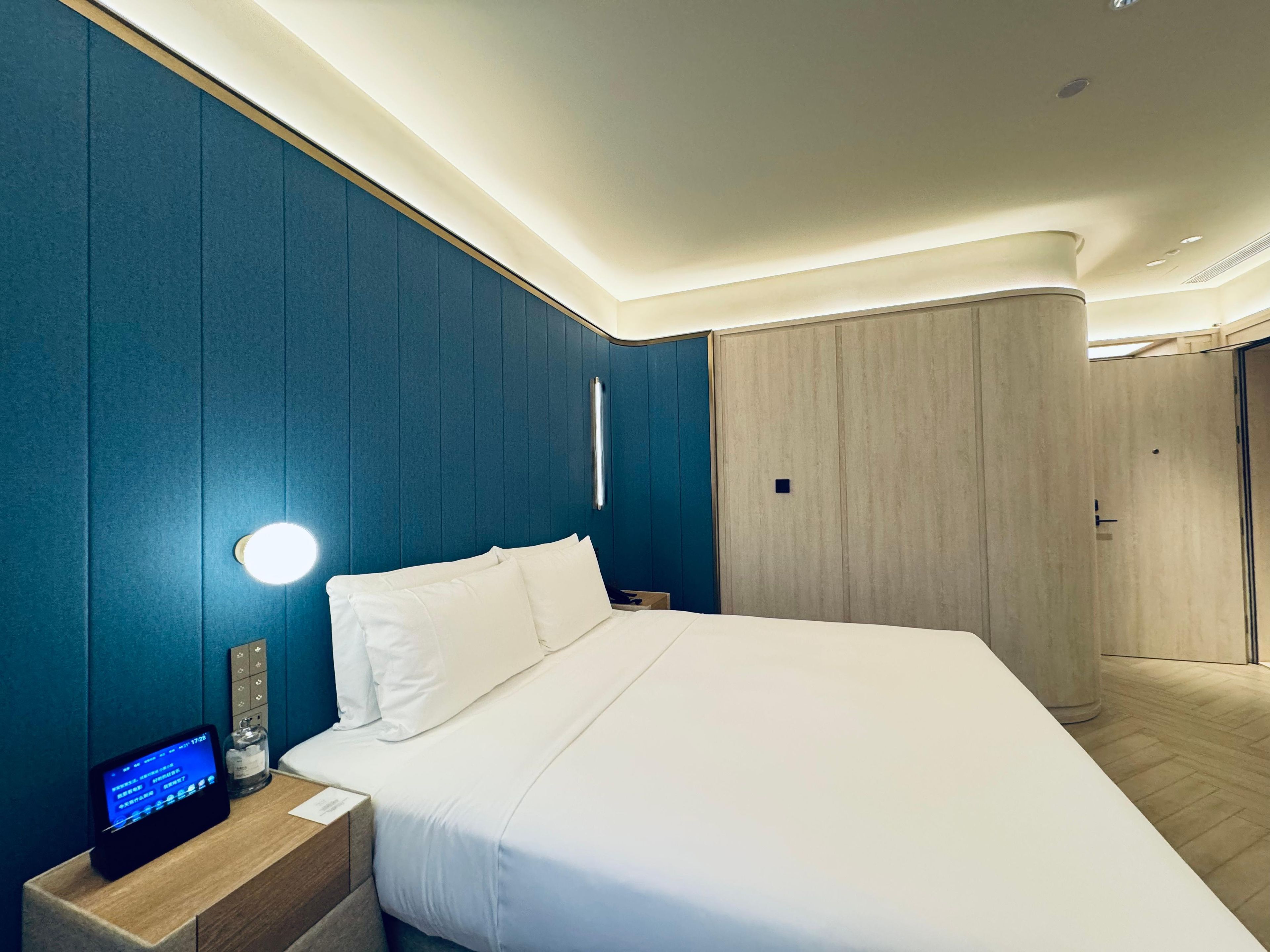 A modern hotel room with a large bed, a bedside table with a digital device, and a wardrobe.