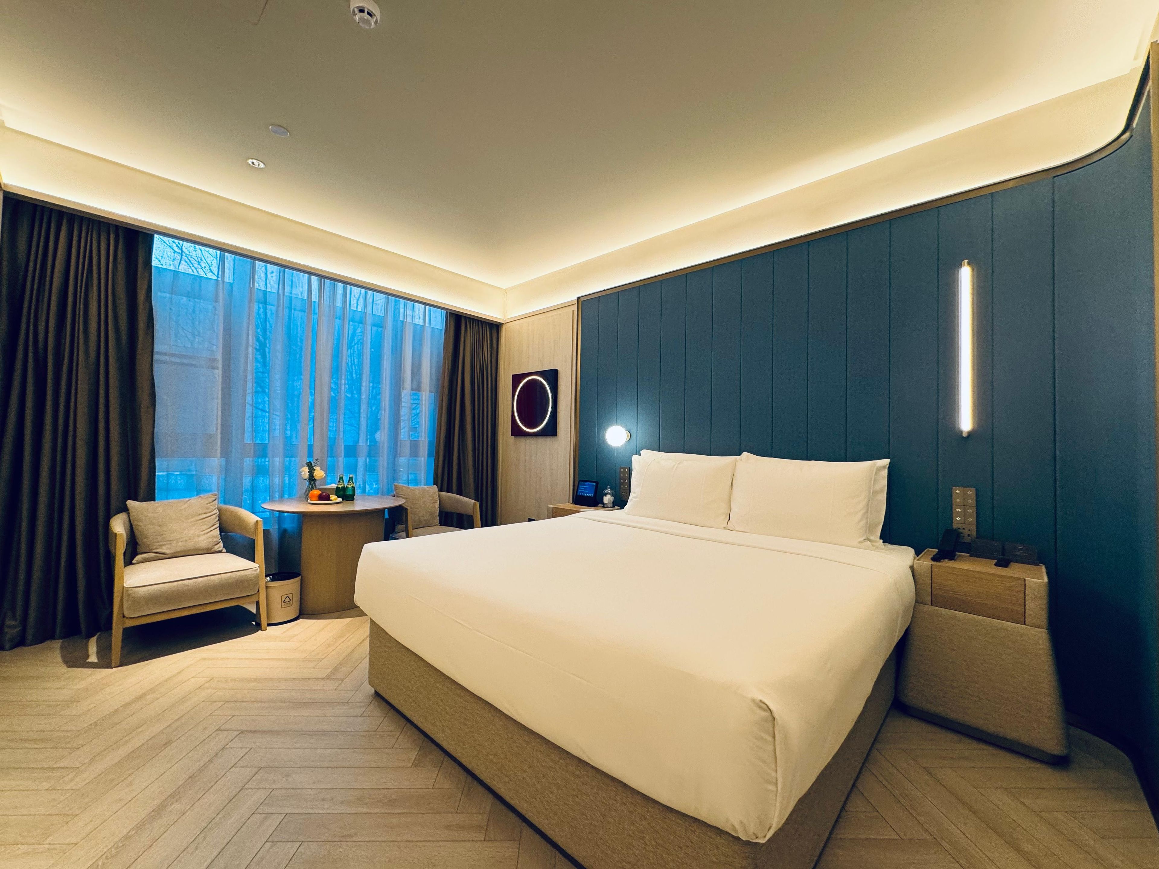 A modern hotel room with a large bed, a small table with a chair, a bedside table, and a view of the outdoors.