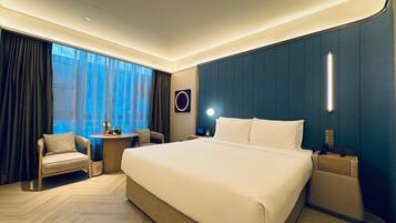 A modern hotel room with a large bed, a small table with a chair, a bedside table, and a view of the outdoors.