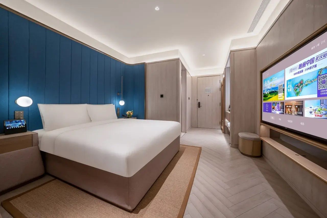 A modern hotel room with a large bed, bedside tables, a TV, and a wall-mounted screen displaying information.