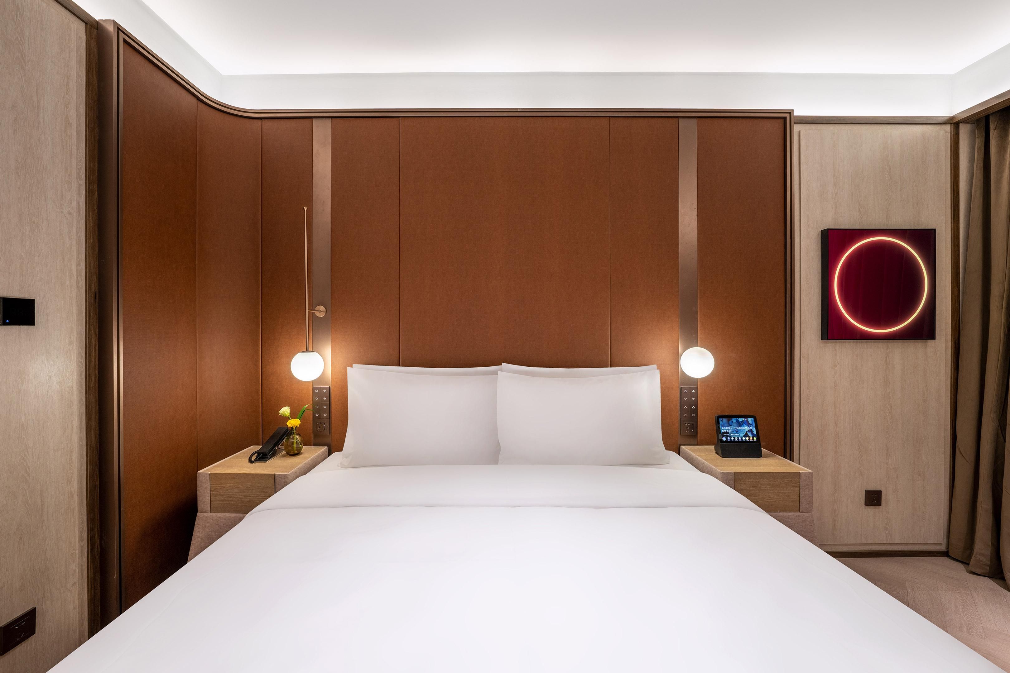 Quarto de hotel com uma cama grande, duas mesas de cabeceira, um telefone fixado na parede, um relógio digital e uma luminária circular.