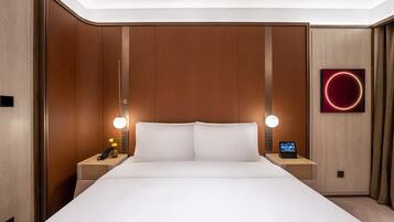 Quarto de hotel com uma cama grande, duas mesas de cabeceira, um telefone fixado na parede, um relógio digital e uma luminária circular.
