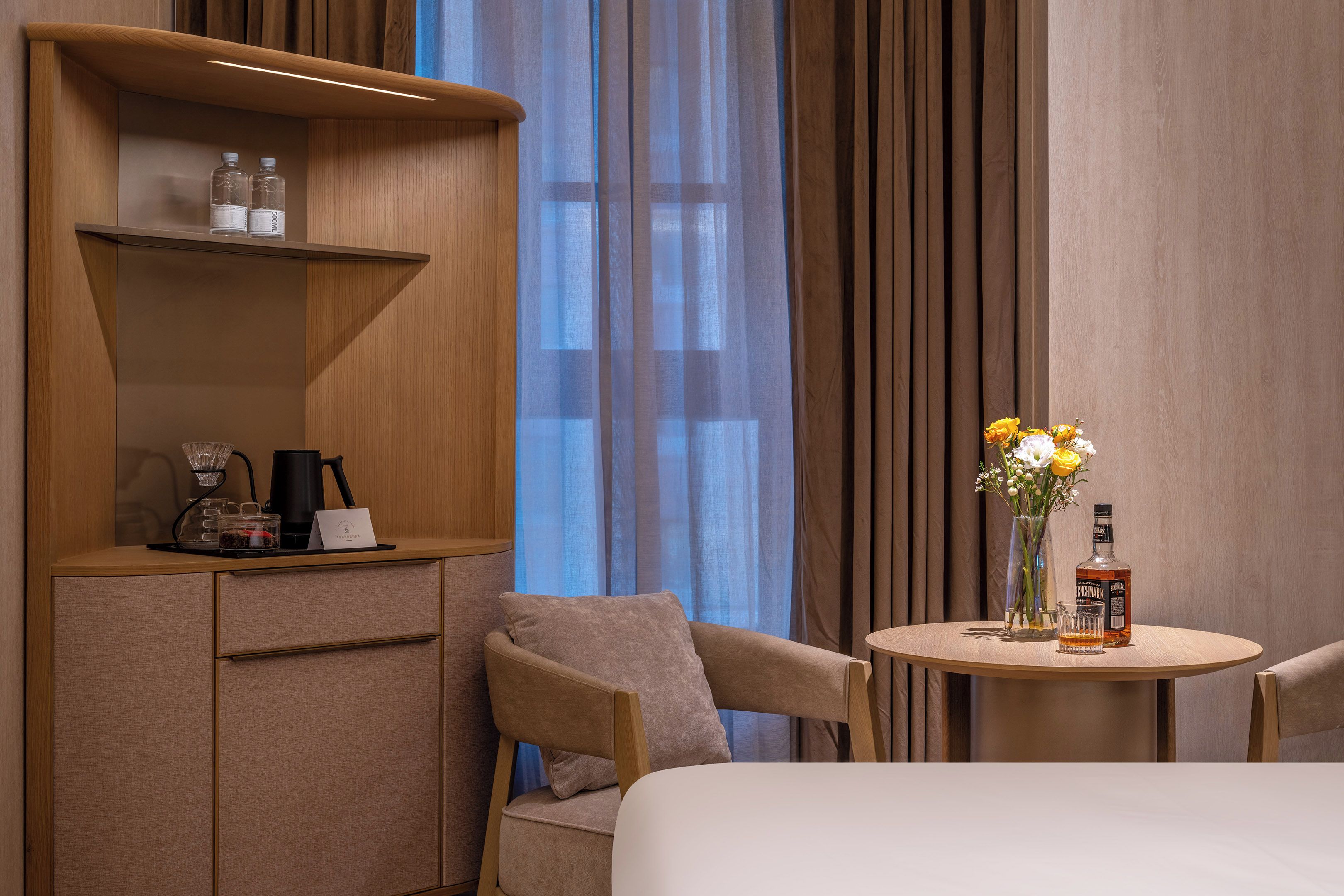Quarto de hotel com uma cama, uma escrivaninha com cafeteira, uma garrafa e um vaso com flores.