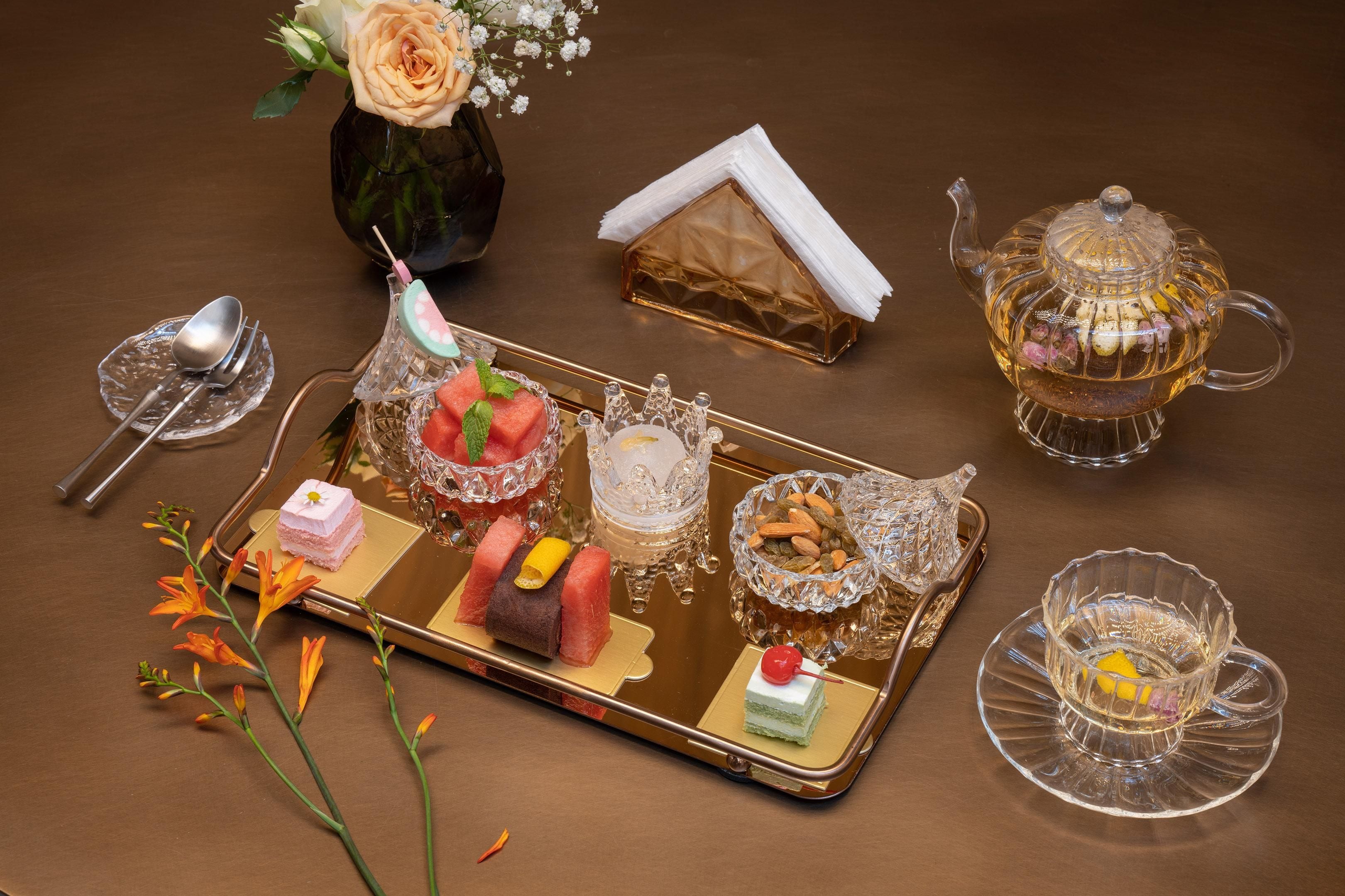A tray with various desserts, a teapot, and a vase with flowers on a table.