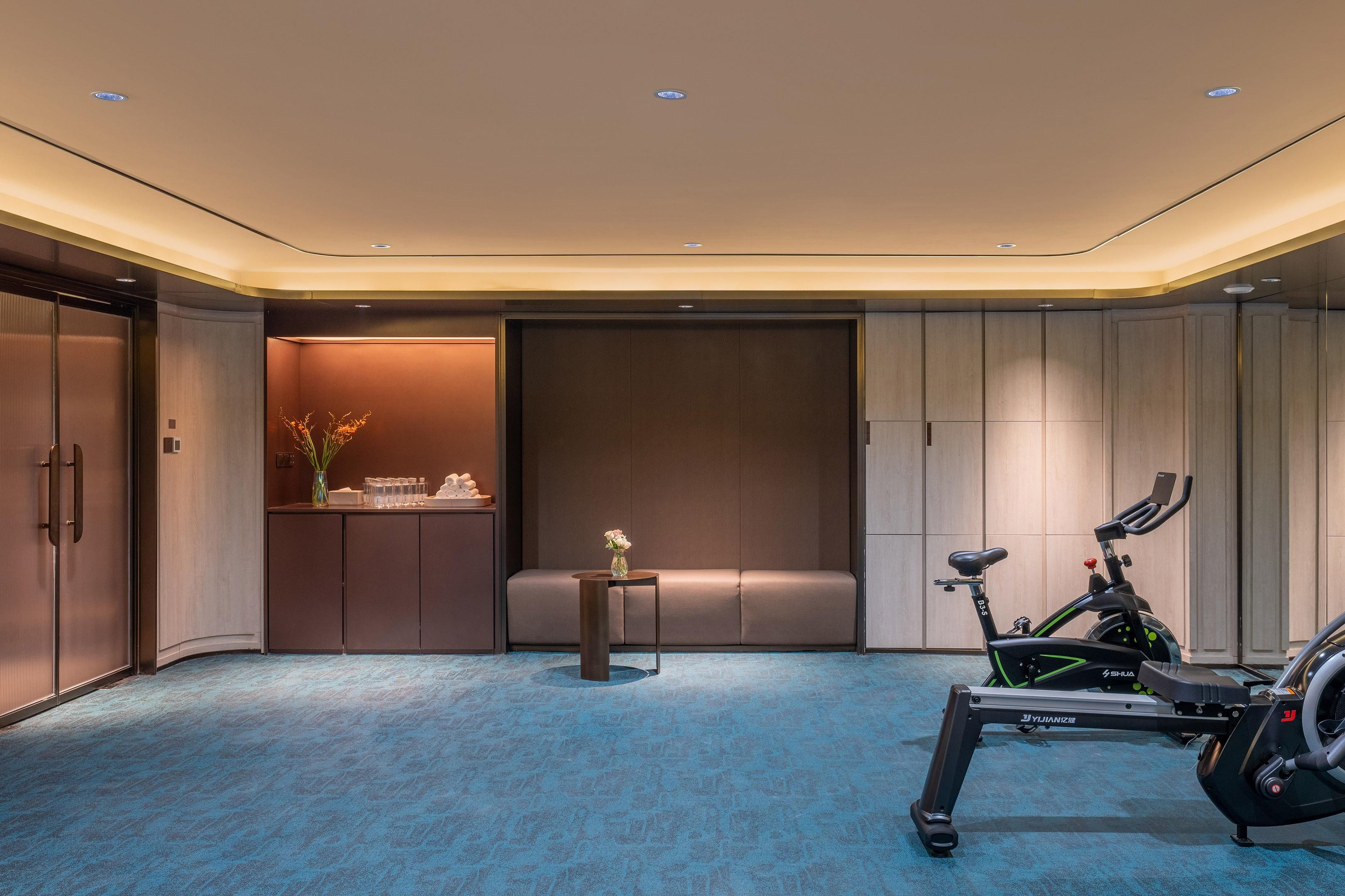 Quarto de hotel moderno com bicicleta ergométrica, uma pequena mesa com um vaso e um espaço de estar.