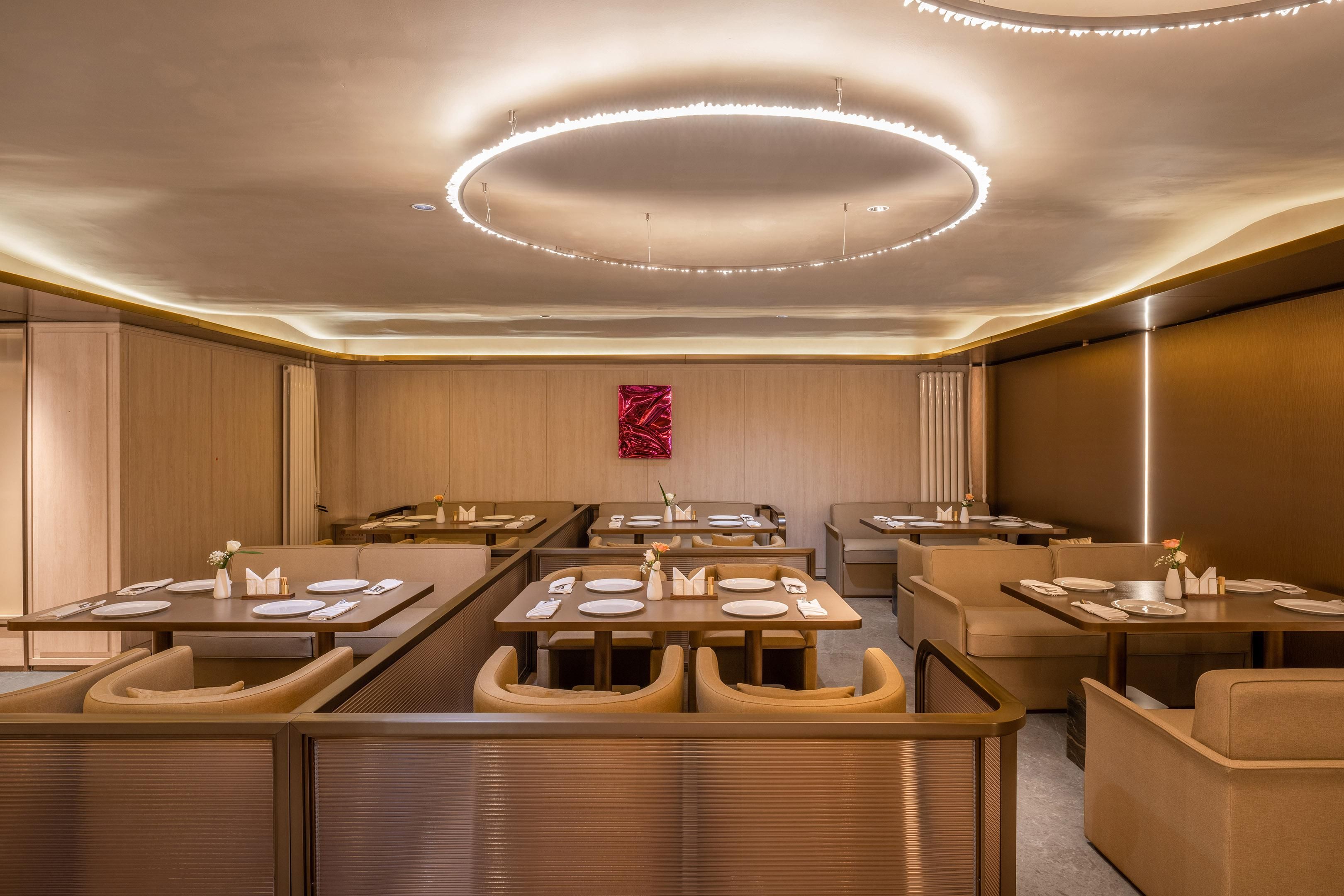 A modern hotel dining area with multiple tables and booths, a central light fixture, and a decorative wall piece.