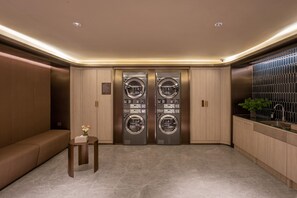 A modern, well-lit laundry room with two large, stainless steel front-loading washers and dryers, a wooden bench, and a small table with a vase of flowers. - Crystal Orange Hotel Beijing Yizhuang JD (Beijing)