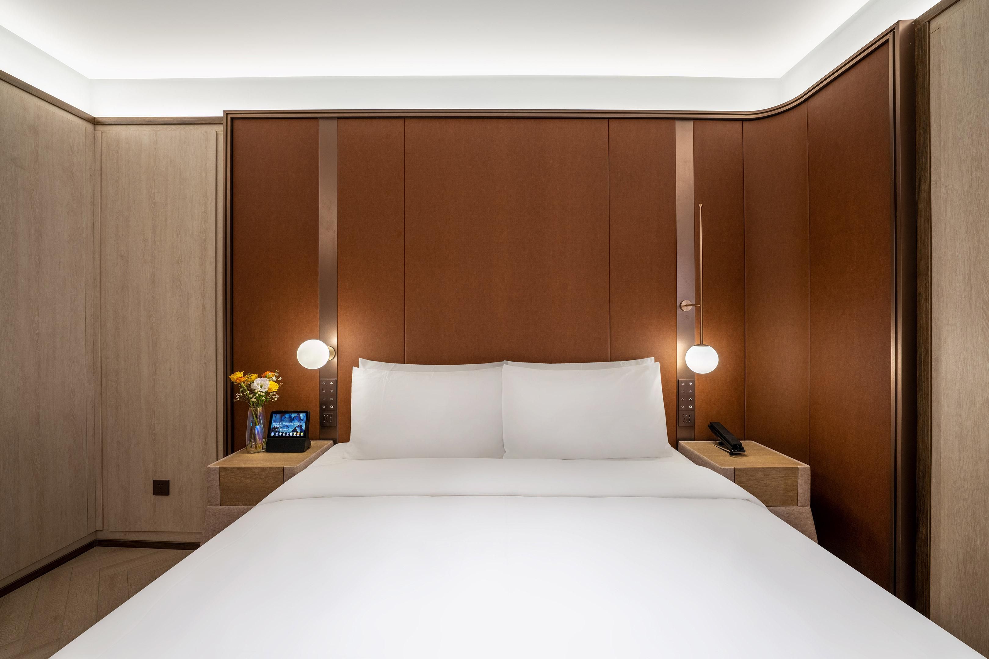 Quarto de hotel com uma cama grande, paredes de painel de madeira, mesa de cabeceira com telefone e relógio digital.