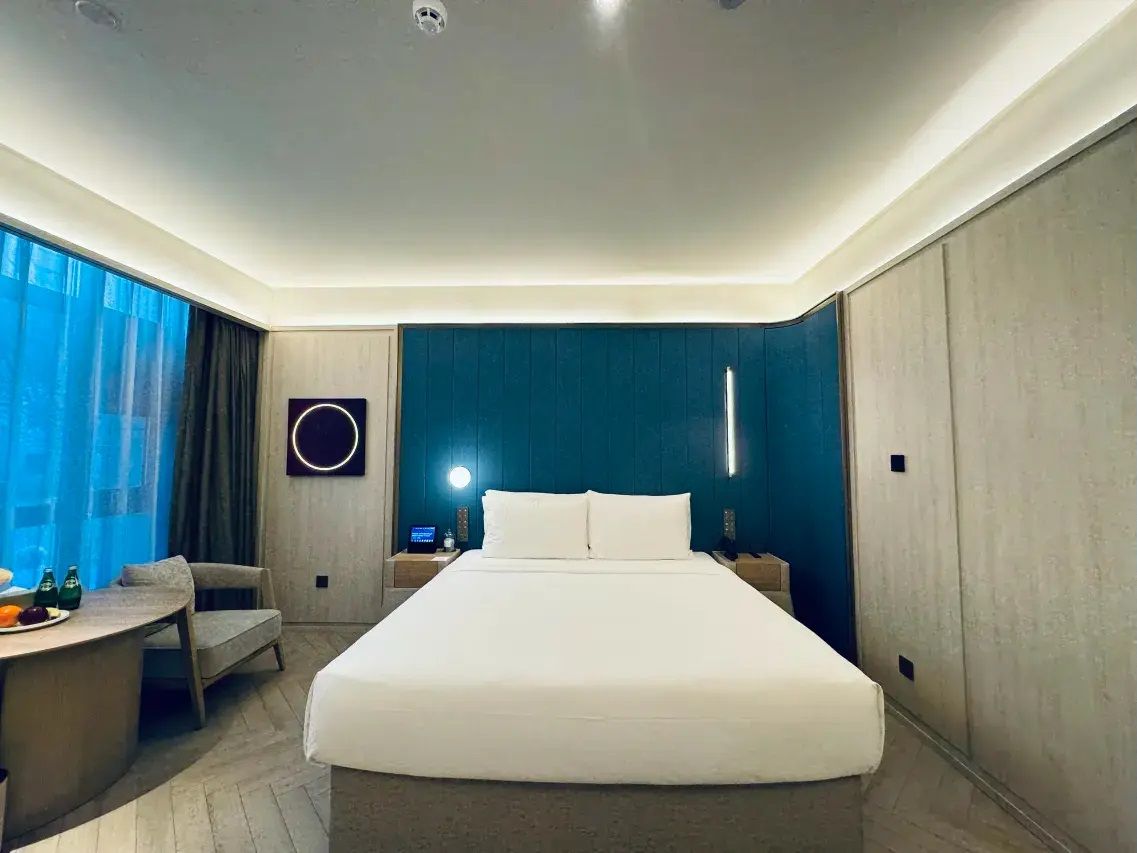 Quarto de hotel moderno com uma cama grande, uma escrivaninha com cadeira, uma mesa redonda com frutas e vista para a cidade através de grandes janelas.