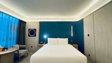 Quarto de hotel moderno com uma cama grande, uma escrivaninha com cadeira, uma mesa redonda com frutas e vista para a cidade através de grandes janelas.