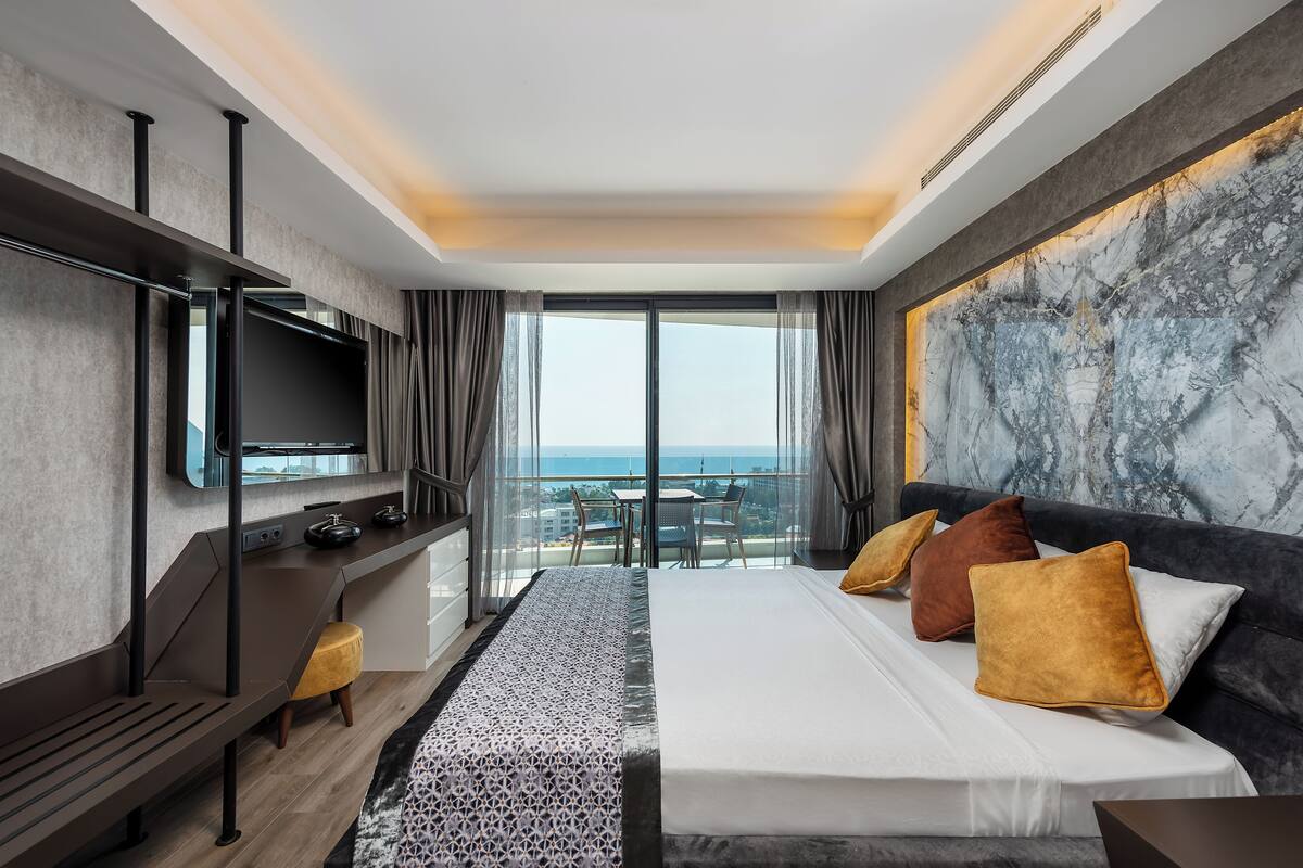 A modern hotel room with a large bed, a desk, a flat-screen TV, and a balcony with a view of the sea.