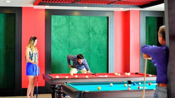 Una mesa de billar en una habitación con paredes verdes y rojas, un techo con iluminación roja, y una persona jugando al billar.