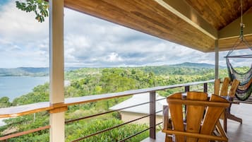 Terraza de madera con vistas a un lago y bosque. Hay dos sillas de madera y una hamaca.