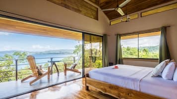 Un dormitorio con una cama grande, vistas al mar y un balcón con sillas de madera.