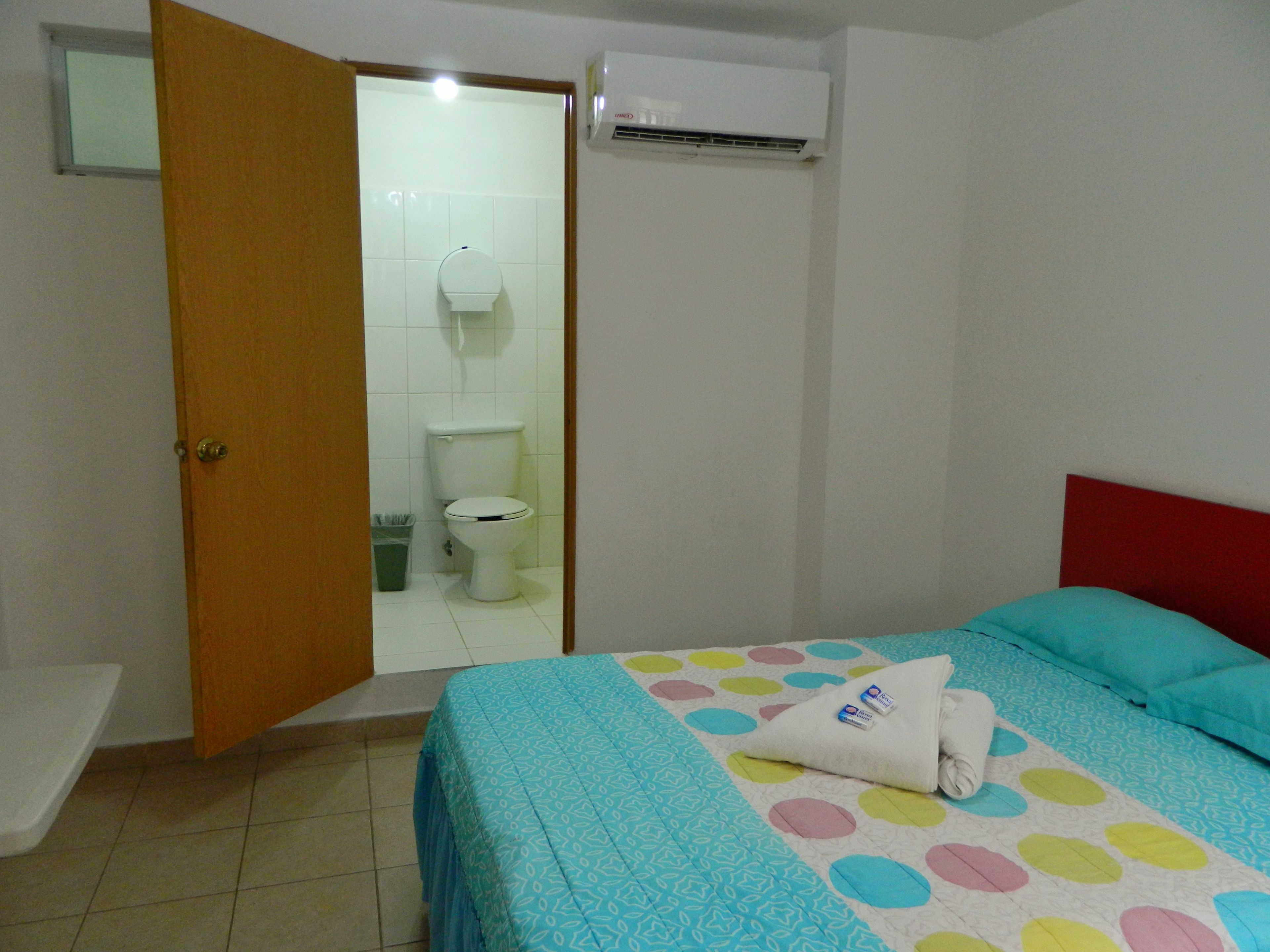 Un dormitorio con cama, cabecera roja, baño con inodoro y ducha, y un equipo de aire acondicionado.