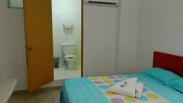 Un dormitorio con cama, cabecera roja, baño con inodoro y ducha, y un equipo de aire acondicionado.