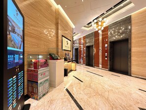 Un hall de hotel con mostrador de recepción, pantalla digital, suelo de mármol, ascensores y un candelabro.