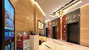 Un hall de hotel con mostrador de recepción, pantalla digital, suelo de mármol, ascensores y un candelabro.