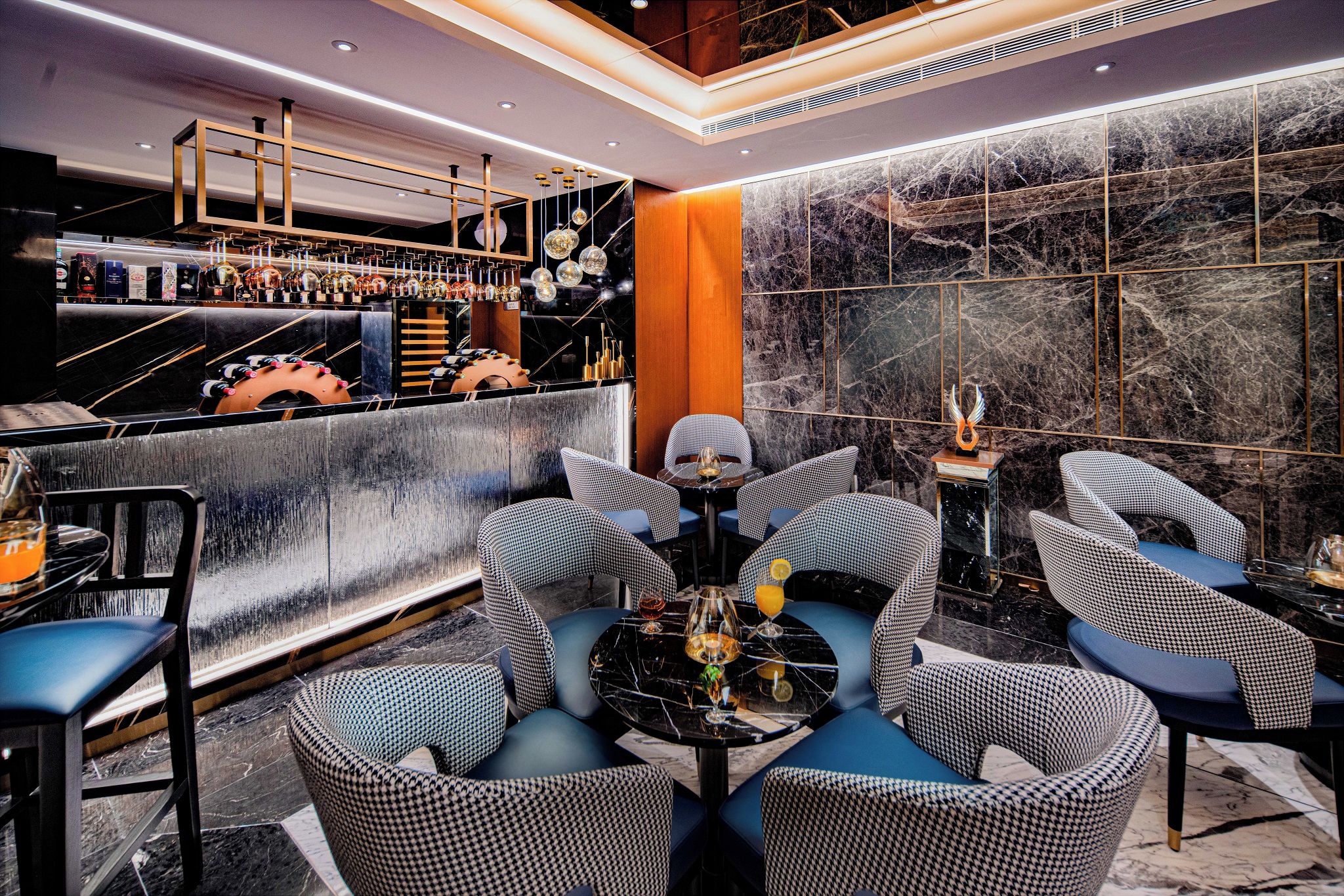 A bar area with a marble wall, a bar counter with various bottles, and seating with a round table.