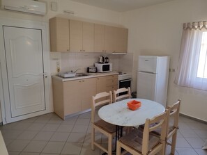 Une cuisine compacte avec un réfrigérateur blanc, un four à micro-ondes, une cuisinière et un évier. On y trouve une table ronde avec des chaises, et un petit saladier orange posé dessus.