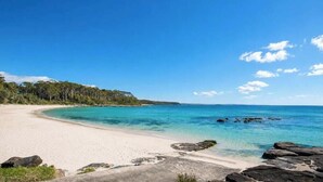 En strand med krystalklart turkis vand, sandet bredde og et skovområde i baggrunden.