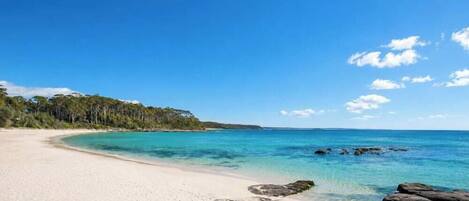 A beach with clear turquoise water, a sandy shore, and a forested area in the background.