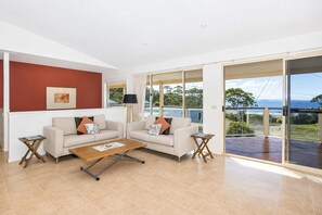 A modern living room with two sofas, a coffee table, and a large sliding glass door leading to a balcony with a view of the sea.