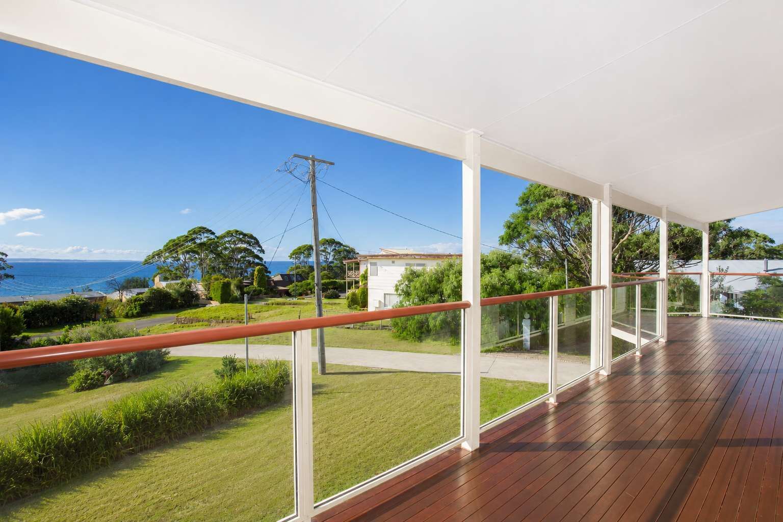 A spacious balcony with a wooden floor and glass railings, overlooking a grassy area and a view of the sea.