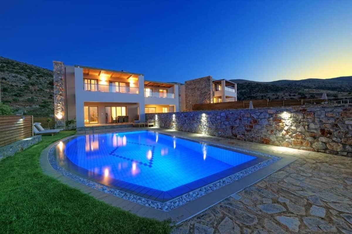 A modern two-story house with a swimming pool, stone walls, and a view of the hills.
