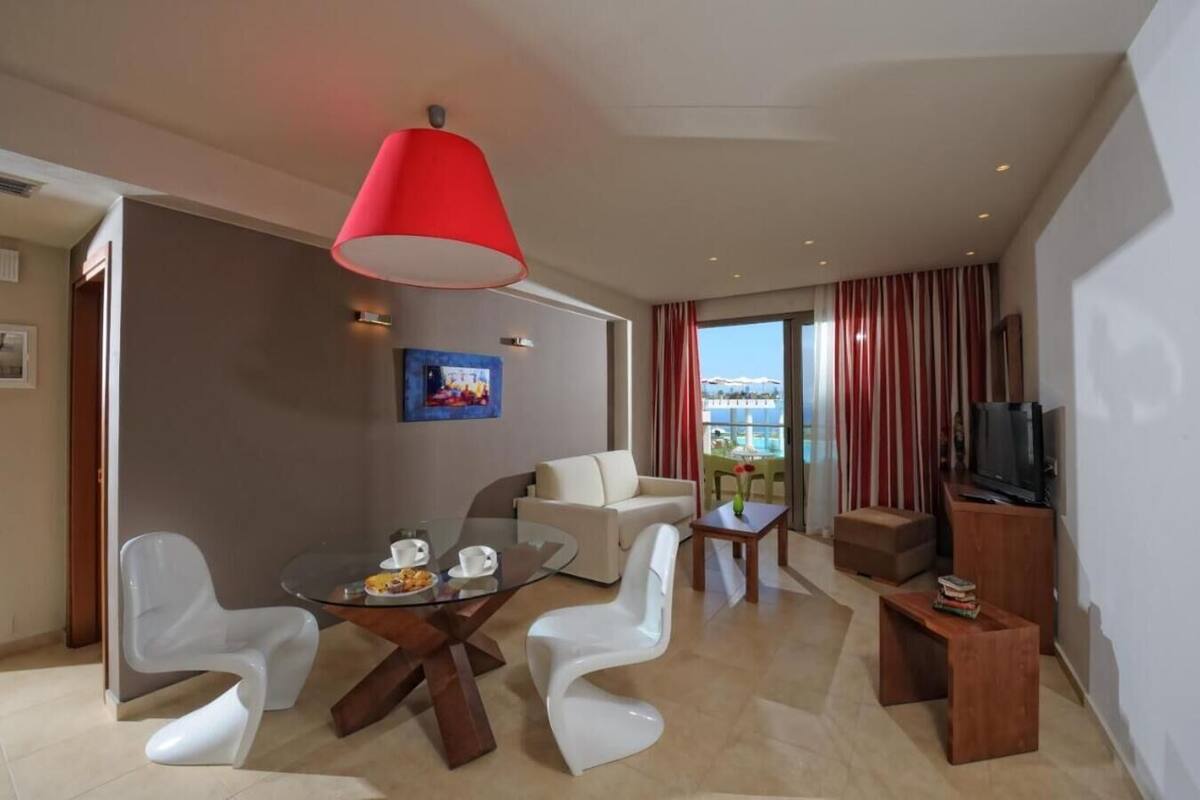A modern hotel room with a glass dining table, white chairs, a sofa, a TV, and a balcony with a view.