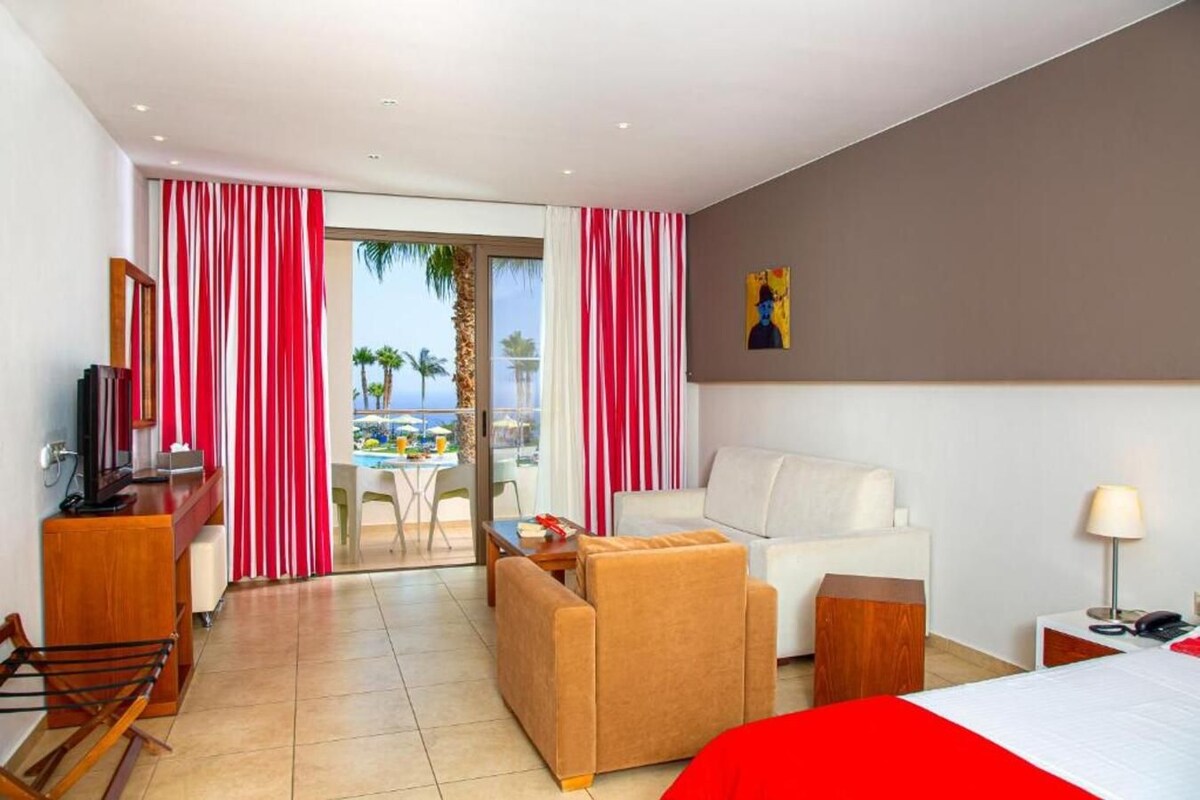 A hotel room with a bed, a sofa, a desk with a TV, a balcony with a pool view, and a painting on the wall.