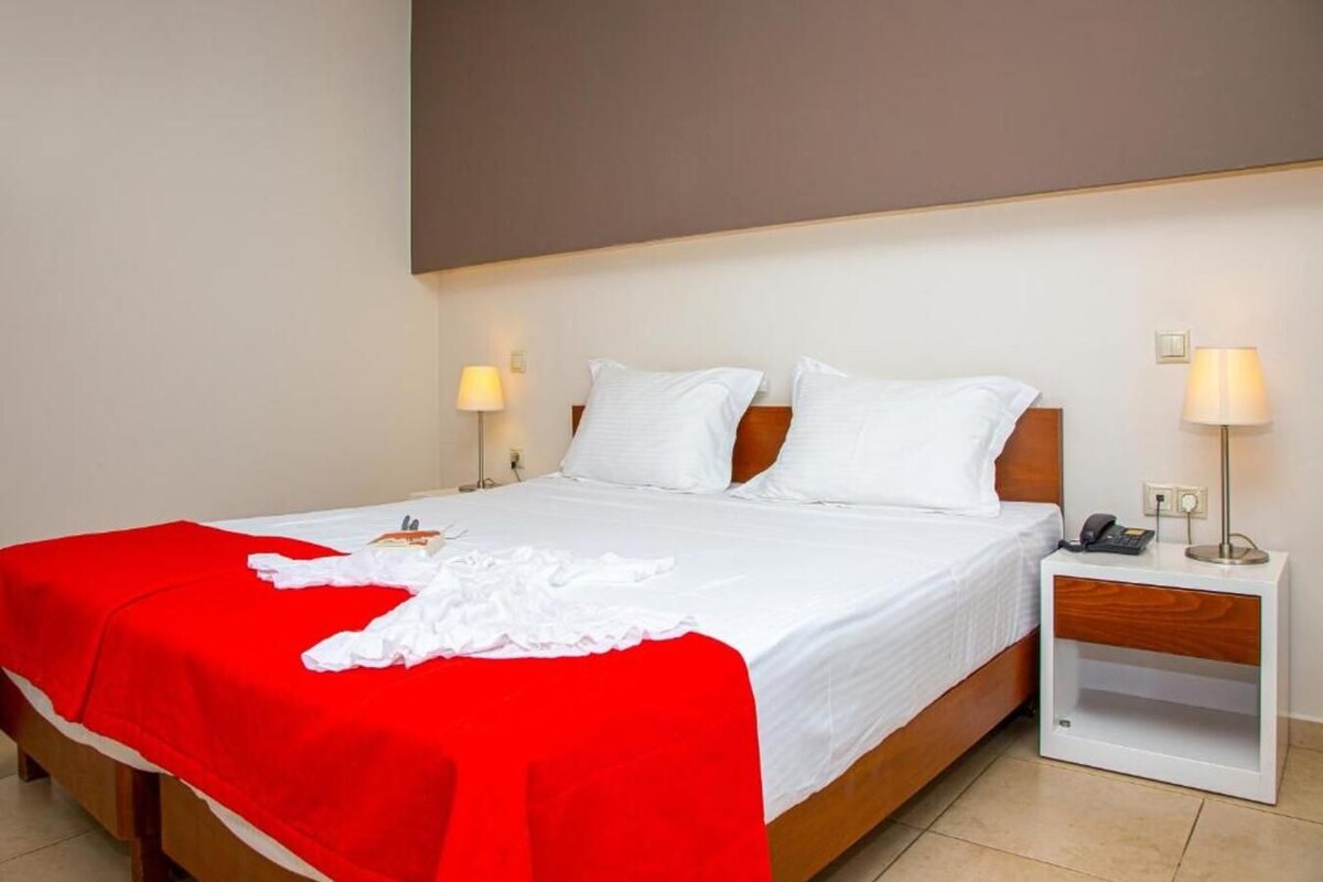 A hotel room with a bed, a red blanket, white pillows, a wooden headboard, bedside lamps, and a nightstand with a phone.