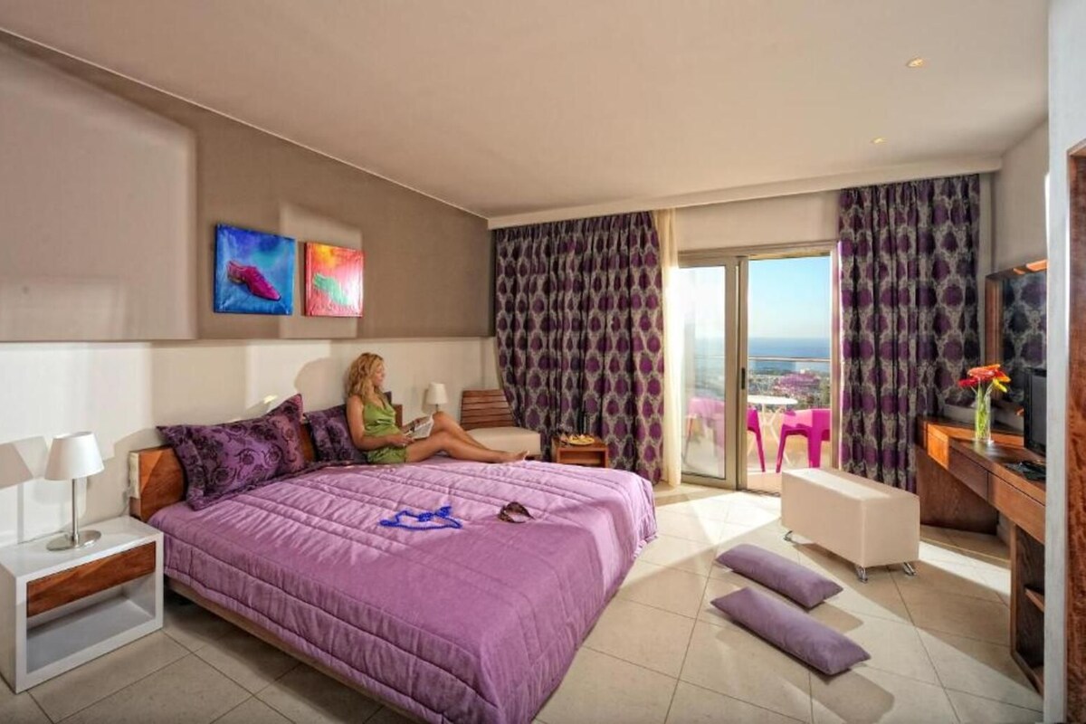 A modern hotel room with a large bed, a balcony with a view, and a desk with a chair.