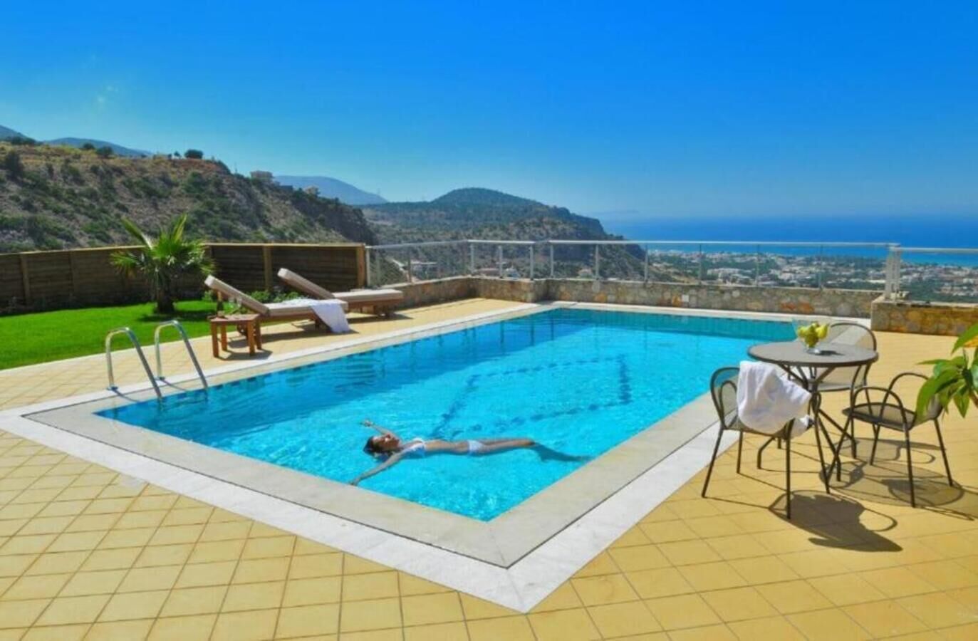 A swimming pool with a view of the ocean and hills, a lounge chair, and a table with chairs.