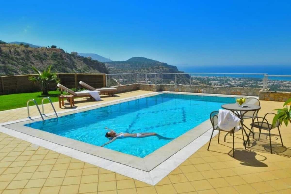 A swimming pool with a view of the ocean and hills, a lounge chair, and a table with chairs.