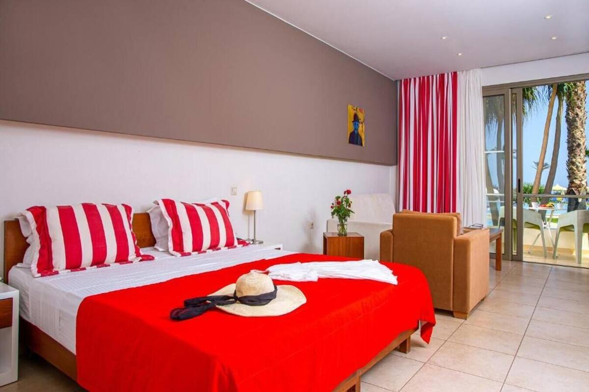 A hotel room with a bed, a chair, a table, and a view of palm trees.