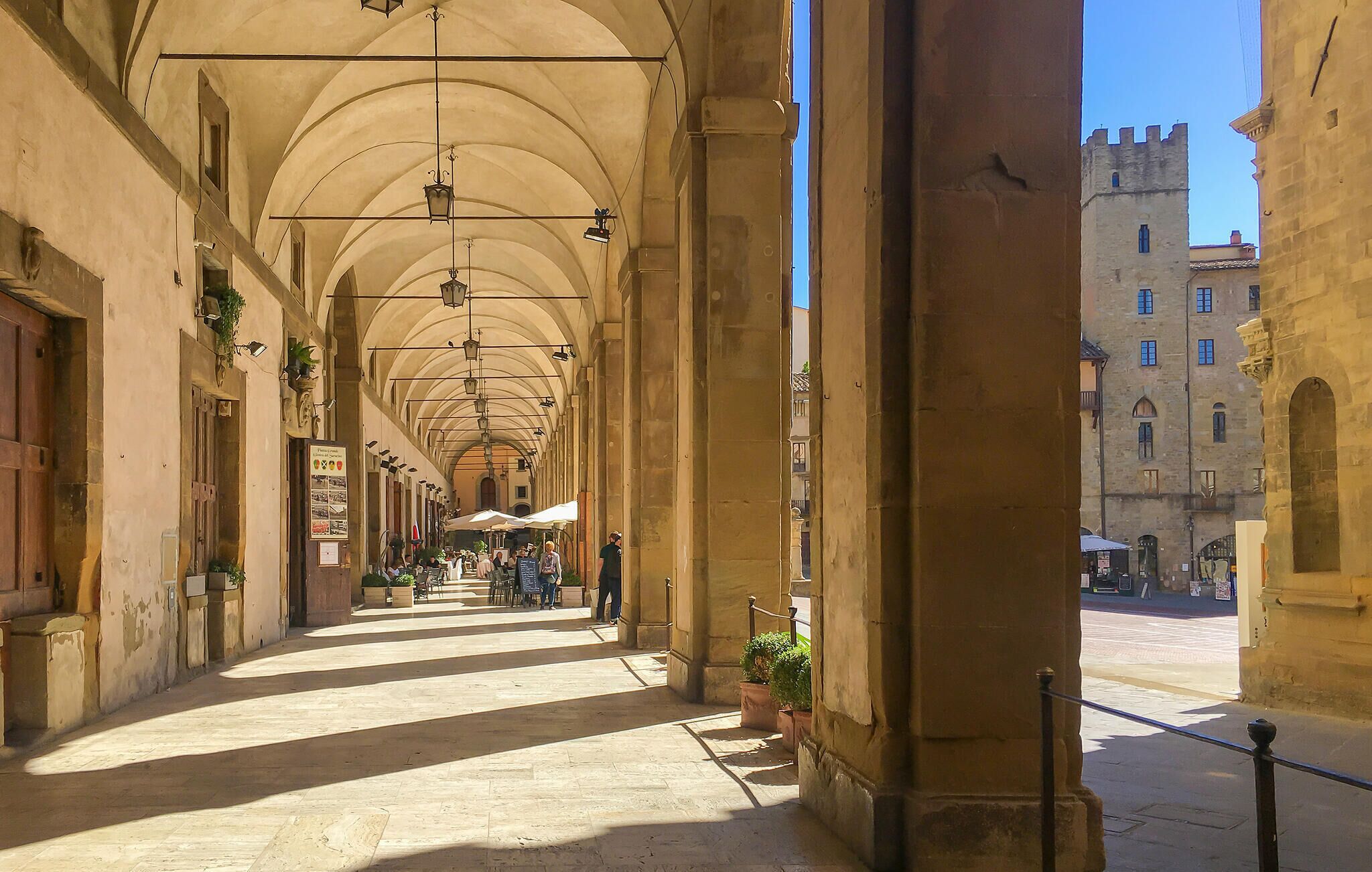 A covered pedestrian walkway with arches, potted plants, and outdoor seating.