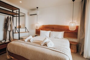 A hotel room with a wooden headboard, a large bed with white linens, two bedside lamps, a wooden side table, and a mirror with clothes hanging. - Sagredo Boutique Hotel And Spa (Kos)