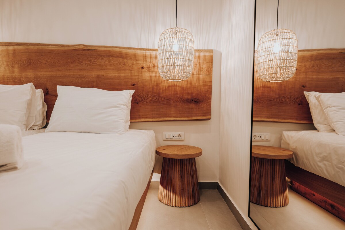 A hotel room with a wooden headboard, a round wooden side table, and a hanging light fixture.