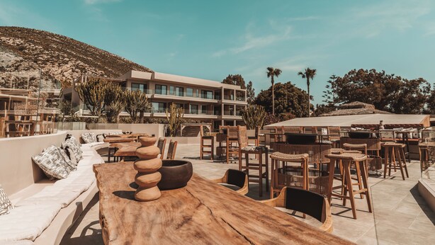 An outdoor seating area with wooden tables and chairs, a sofa with cushions, and a large building in the background. - Sagredo Boutique Hotel And Spa (Kos)