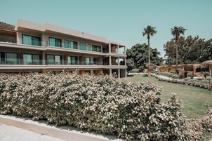 A modern hotel with a multi-story structure, large windows, and a well-maintained garden with hedges and palm trees.