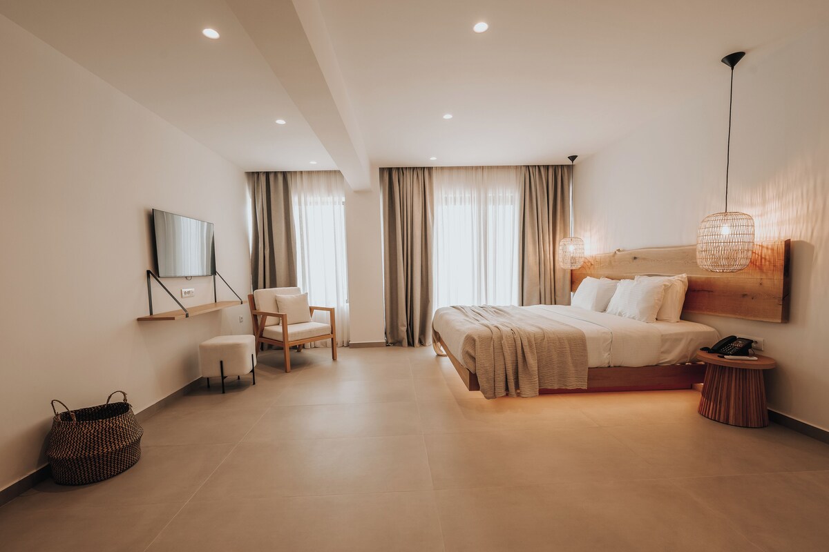 A modern hotel room with a bed, a chair, a small table, a wall-mounted TV, and a hanging light fixture.