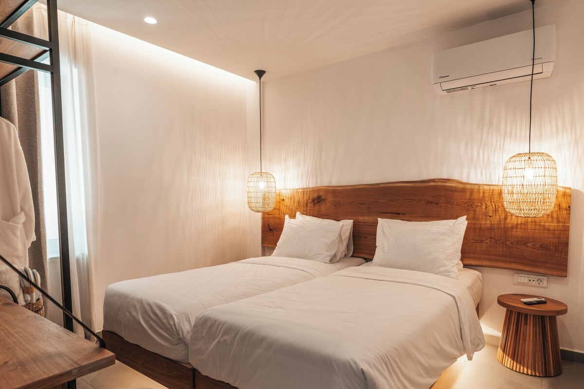 A double bed with a wooden headboard, a bedside table, a round wooden side table, and an air conditioning unit on the wall.