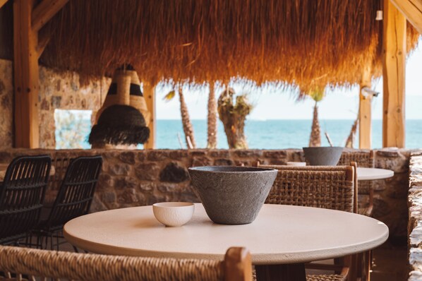 Un restaurant situé en bord de mer, avec des murs en pierre, des meubles en osier et un toit de chaume.