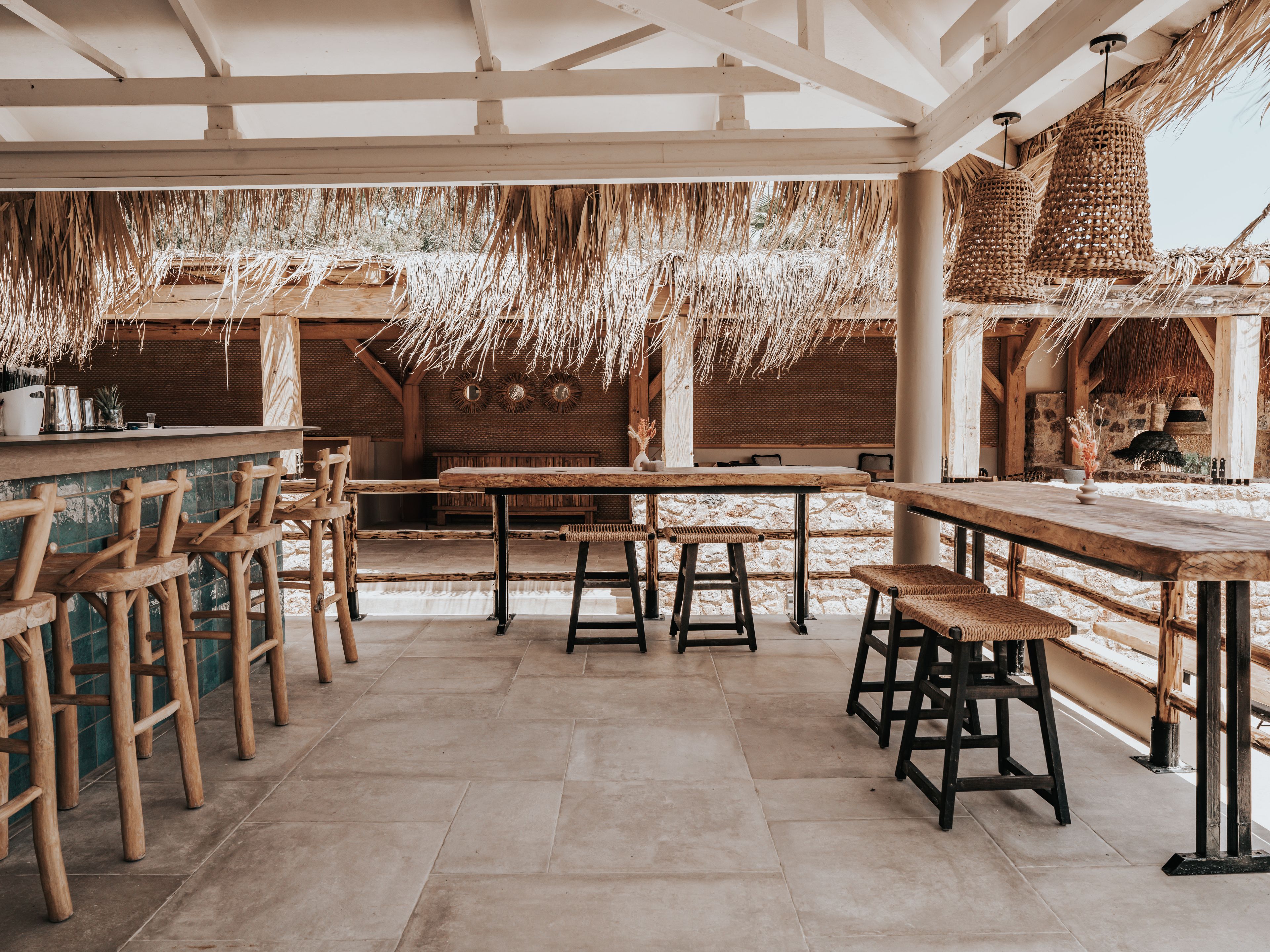 A rustic outdoor dining area with wooden tables and chairs, straw ceiling, and stone walls.