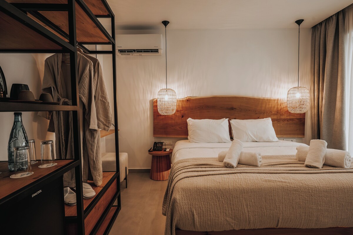 Habitación de hotel con cabecera de madera, cama con sábanas blancas, mesita de noche con lámpara y un armario.